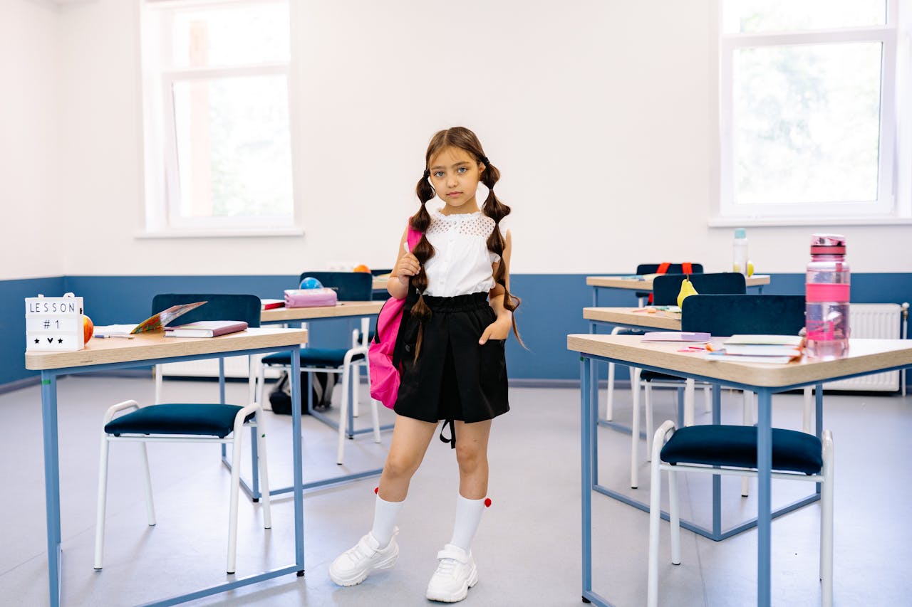A young girl stands confidently in a bright, modern classroom with desks and educational materials.
