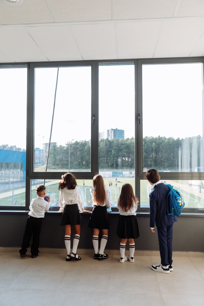 Group of school students in uniforms standing by a large window overlooking a sports field.